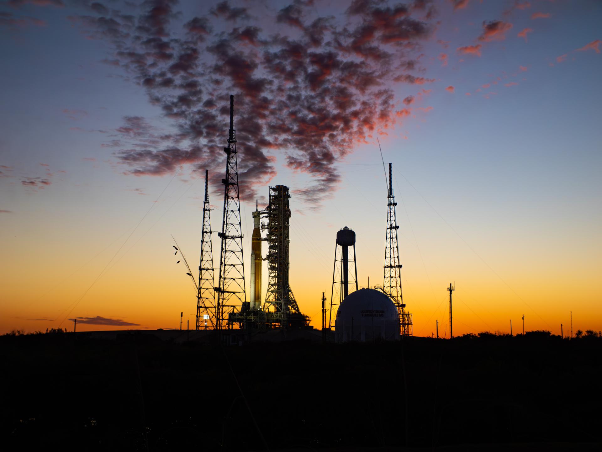 The sun sets behind NASA’s Space Launch System (SLS) rocket and Orion spacecraft as they stand fully assembled atop the mobile launcher at Launch Pad 39B at NASA’s Kennedy Space Center in Florida. The sky glows with warm shades of orange and pink, silhouetting the towering rocket and its solid rocket boosters against the fading light.  Photographed on January 31, 2026, the scene captures teams preparing for a wet dress rehearsal for the Artemis II mission, rehearsing launch countdown timelines and procedures as day turns to night.